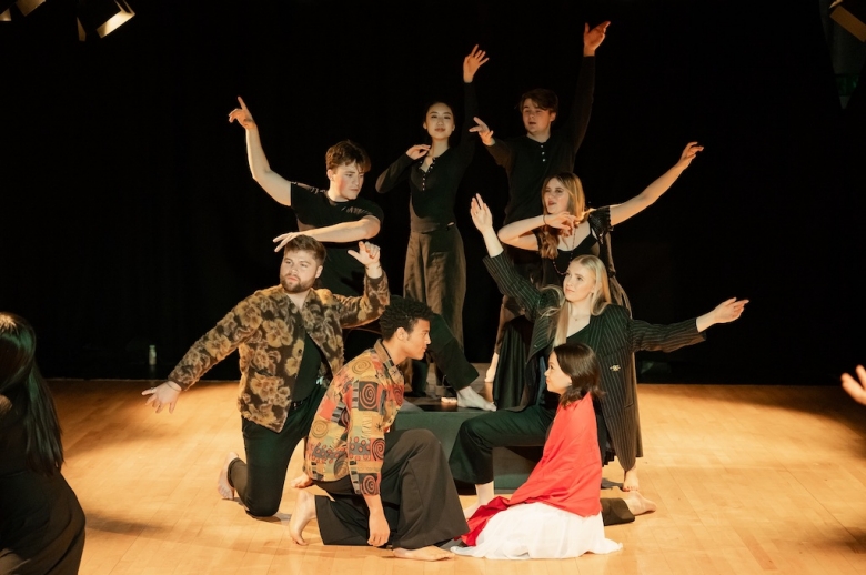 Two actors kneel on stage facing each other while another group of actors with arms outstretched stands or kneels behind them.